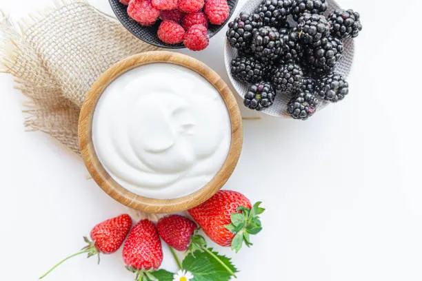 A bowl of white Greek yogurt surrounded by fresh blackberries, blueberries, and strawberries on a white background.